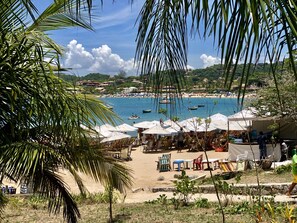 A beach with umbrellas and tables, a view of the sea, and a distant town. - Búzios Summer Park (Búzios)