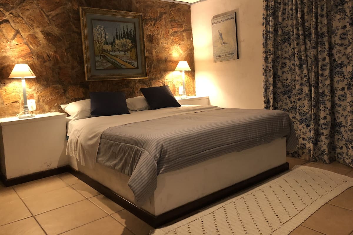 A bedroom with a large bed, two bedside lamps, a painting on the wall, and a floral curtain.
