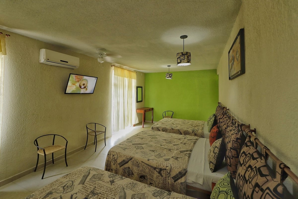 A room with two beds, a chair, a small table, an air conditioner, a ceiling fan, and a TV on the wall.