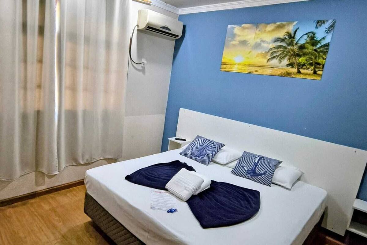 A hotel room with a bed, curtains, an air conditioning unit, a nightstand, and a painting on the wall.