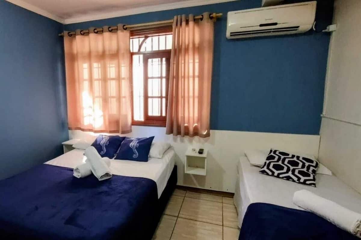 A bedroom with two beds, a window with curtains, an air conditioning unit, and a small bedside table with a phone.