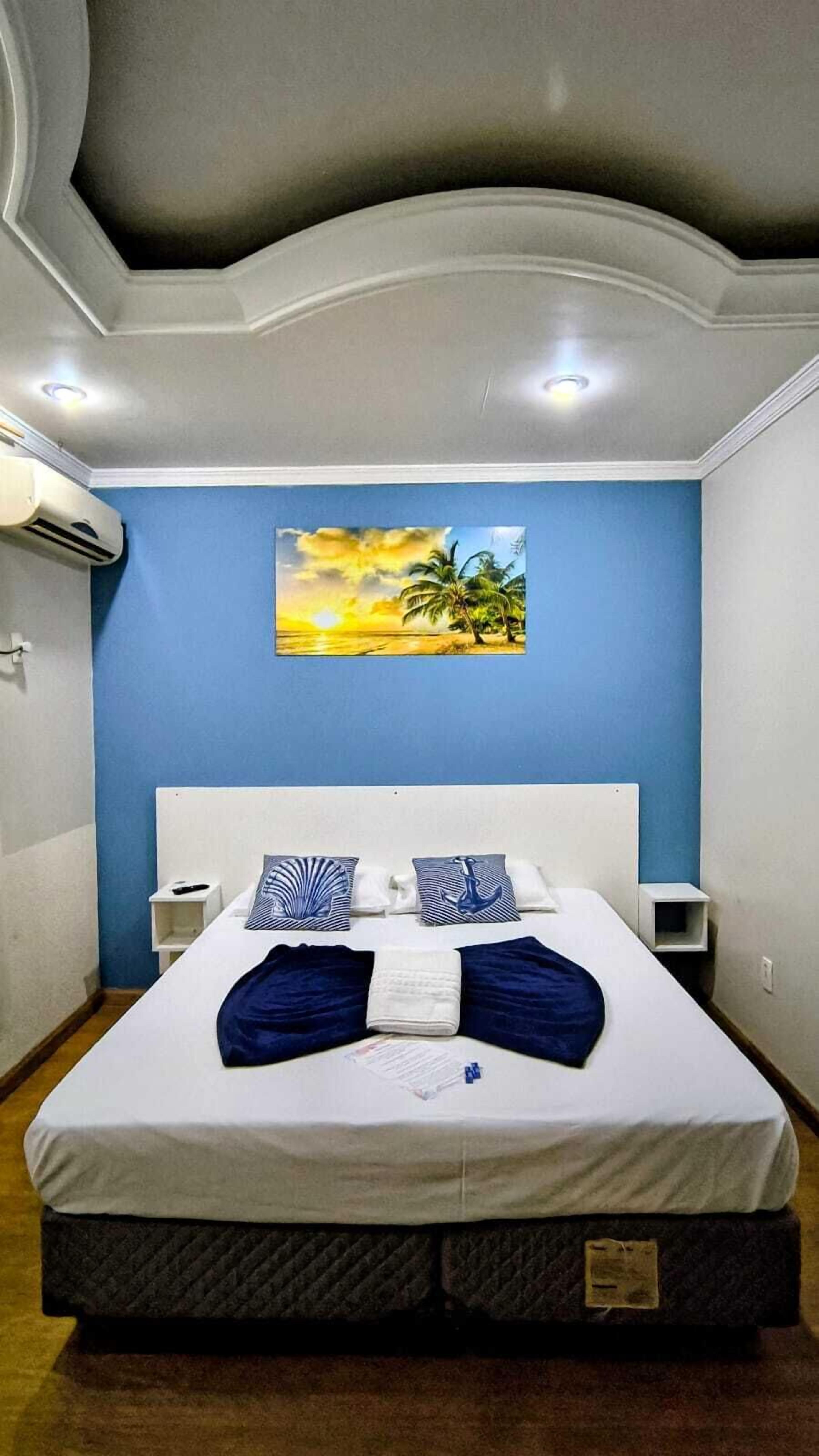 A neatly made bed with a white headboard, blue and white striped pillows, and a blue blanket. There is a painting of a sunset on the wall above the bed.