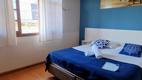 Una cama bien hecha con una colcha azul y almohadas blancas, una ventana con cortinas traslúcidas y un cuadro de una playa en la pared.