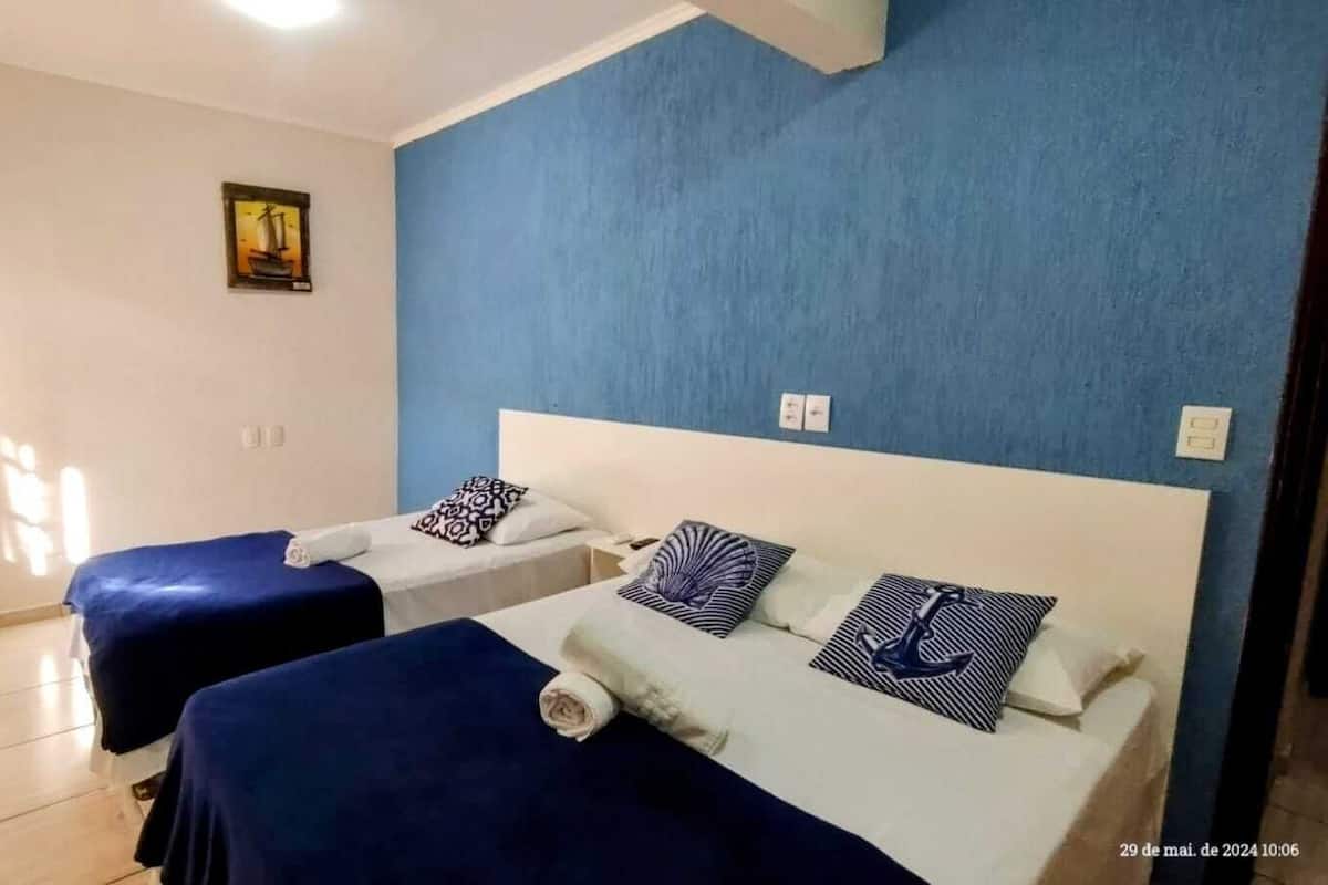 A hotel room with two beds, each with a blue bedspread and matching pillows. There is a framed picture on the wall and a window allowing natural light.
