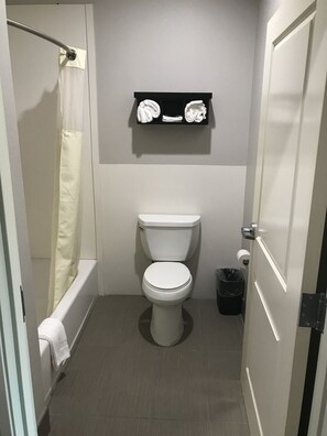 A small, clean bathroom with a toilet, a shower with a white curtain, and a shelf above the toilet. - Grand Villa Inn & Suites Chinatown (Houston)