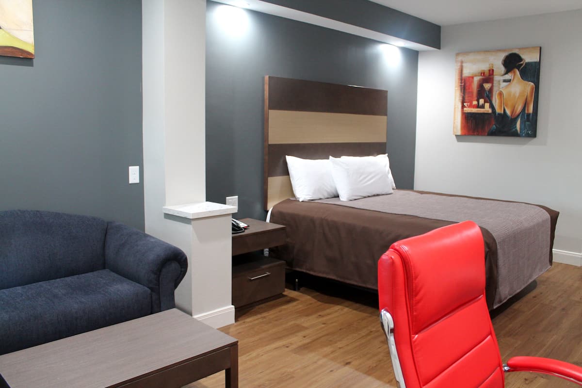 A hotel room with a bed, a red chair, a sofa, a nightstand, and a painting on the wall.