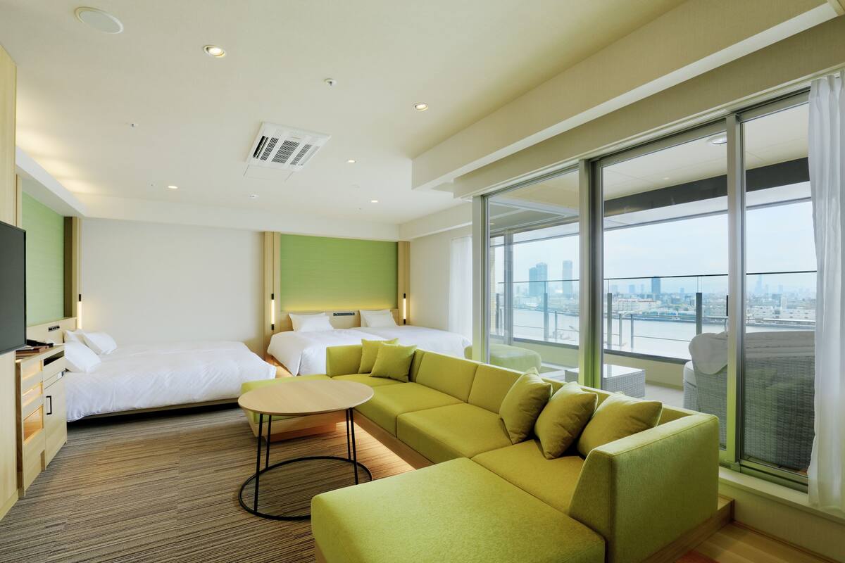 A modern hotel room with a green sofa, a bed, a small round table, and a large window offering a city view.