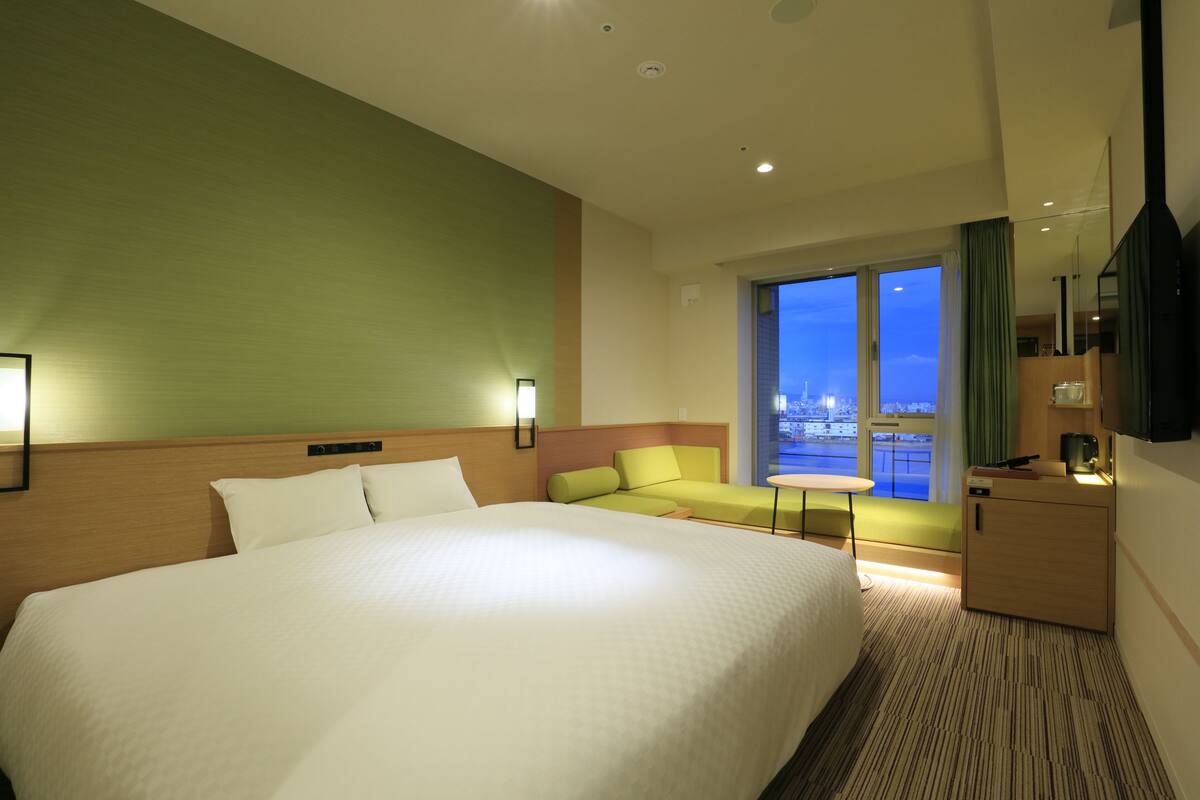 A hotel room with a large bed, a green wall, a sofa, a small table, and a window with a city view.