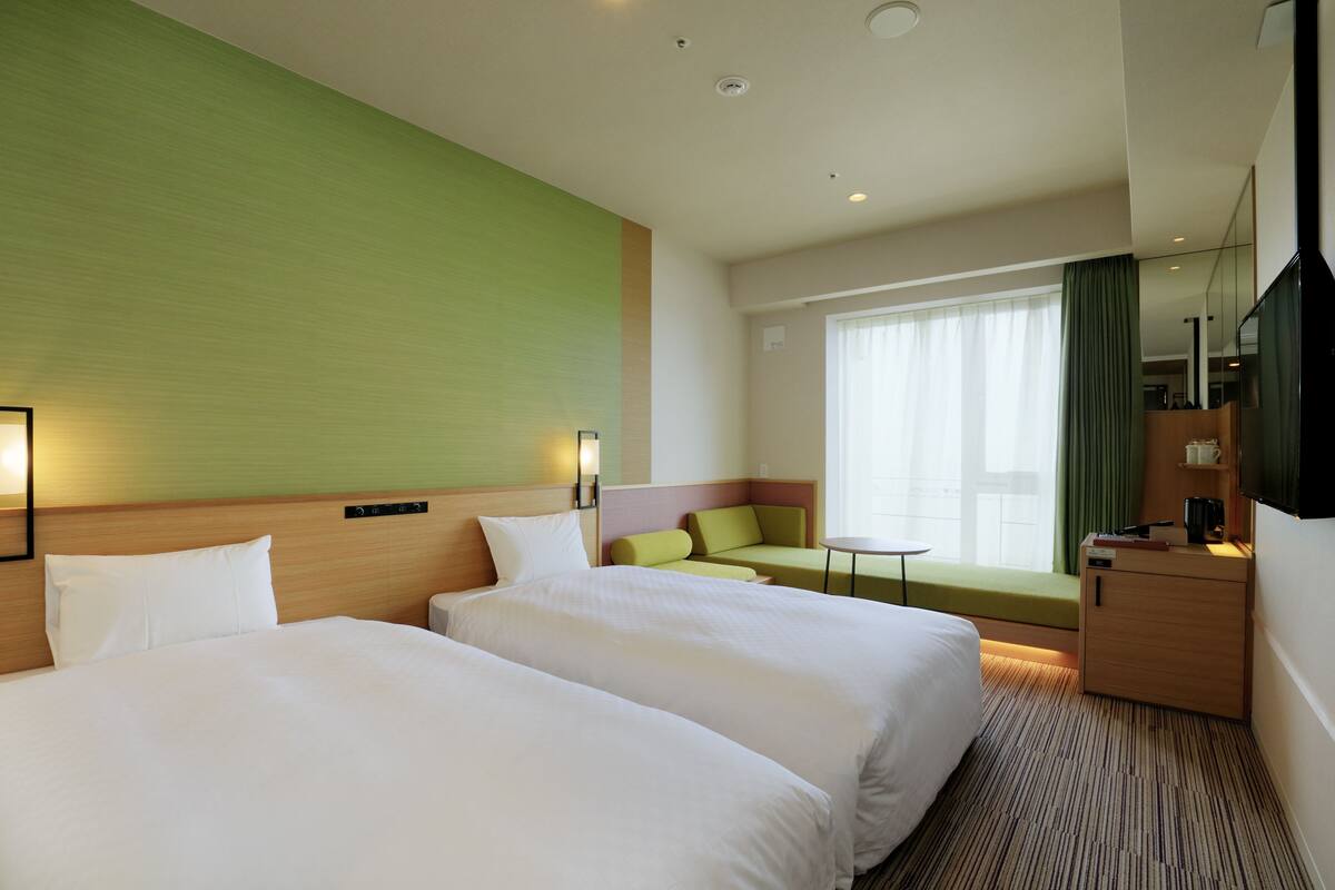 A hotel room with two beds, a green accent wall, a small seating area, a desk, and a television.