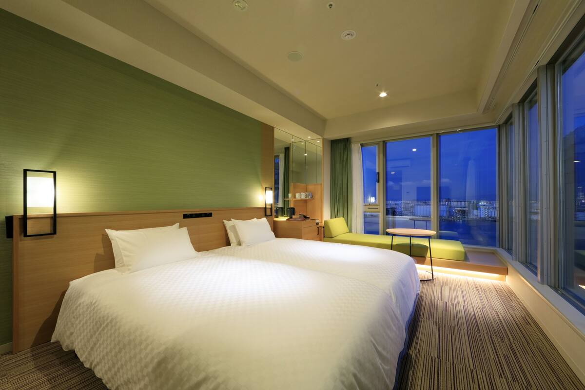 A hotel room with a large bed, a small table, a chair, and a view of the city at night.