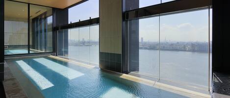 An indoor swimming pool with large windows offering a view of a city skyline and a body of water.
