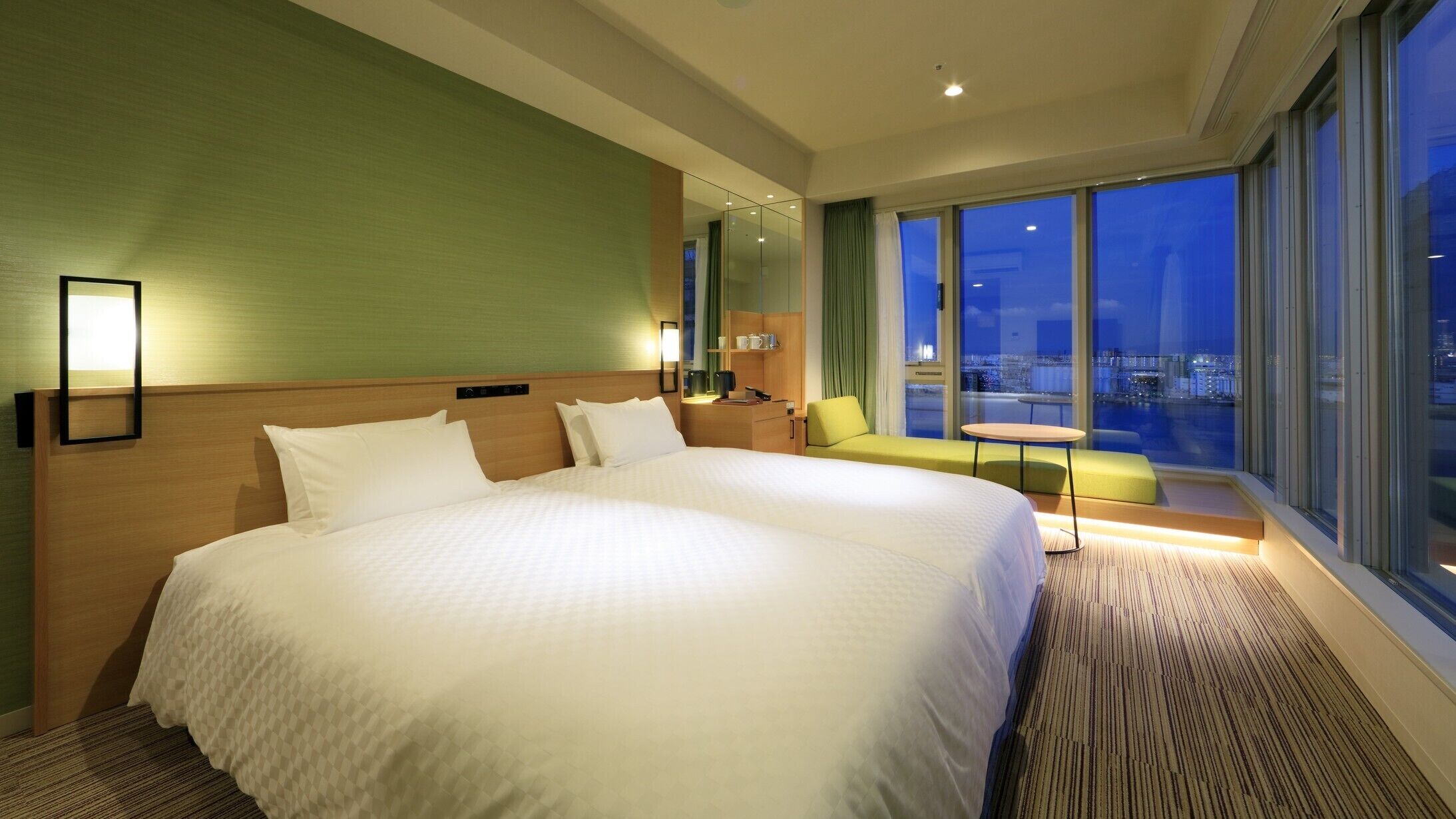 A large bed with a headboard, a bedside table, a chair, a small table, and a view of the city at night.