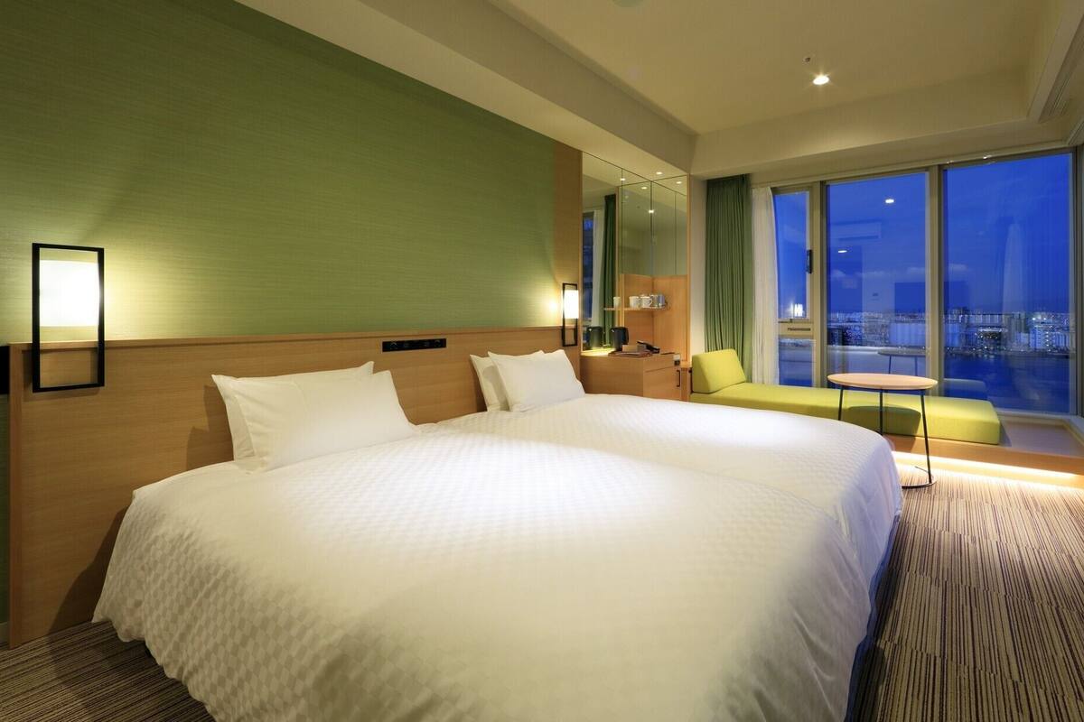 A large bed with a headboard, a bedside table, a chair, a small table, and a view of the city at night.