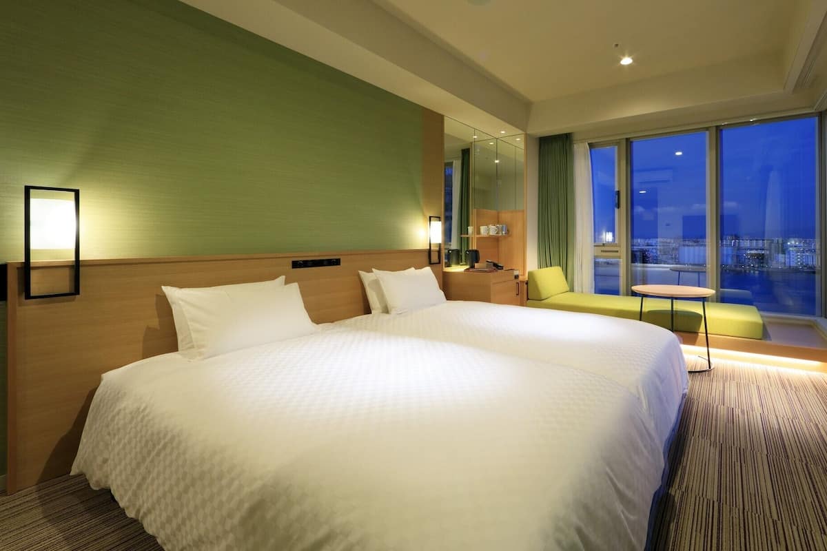 A large bed with a headboard, a bedside table, a chair, a small table, and a view of the city at night.