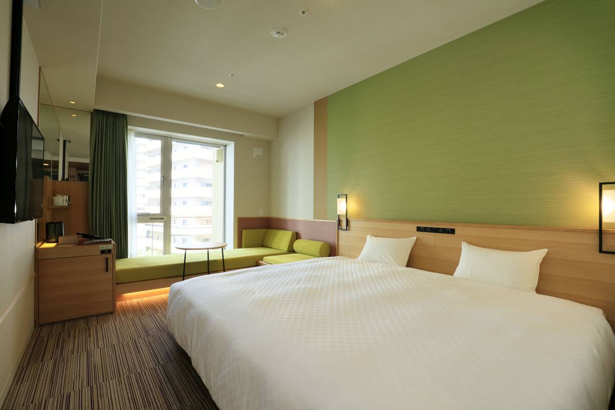A hotel room with a large bed, a seating area with a sofa and armchairs, a desk with a lamp, and a large window with a view of buildings.