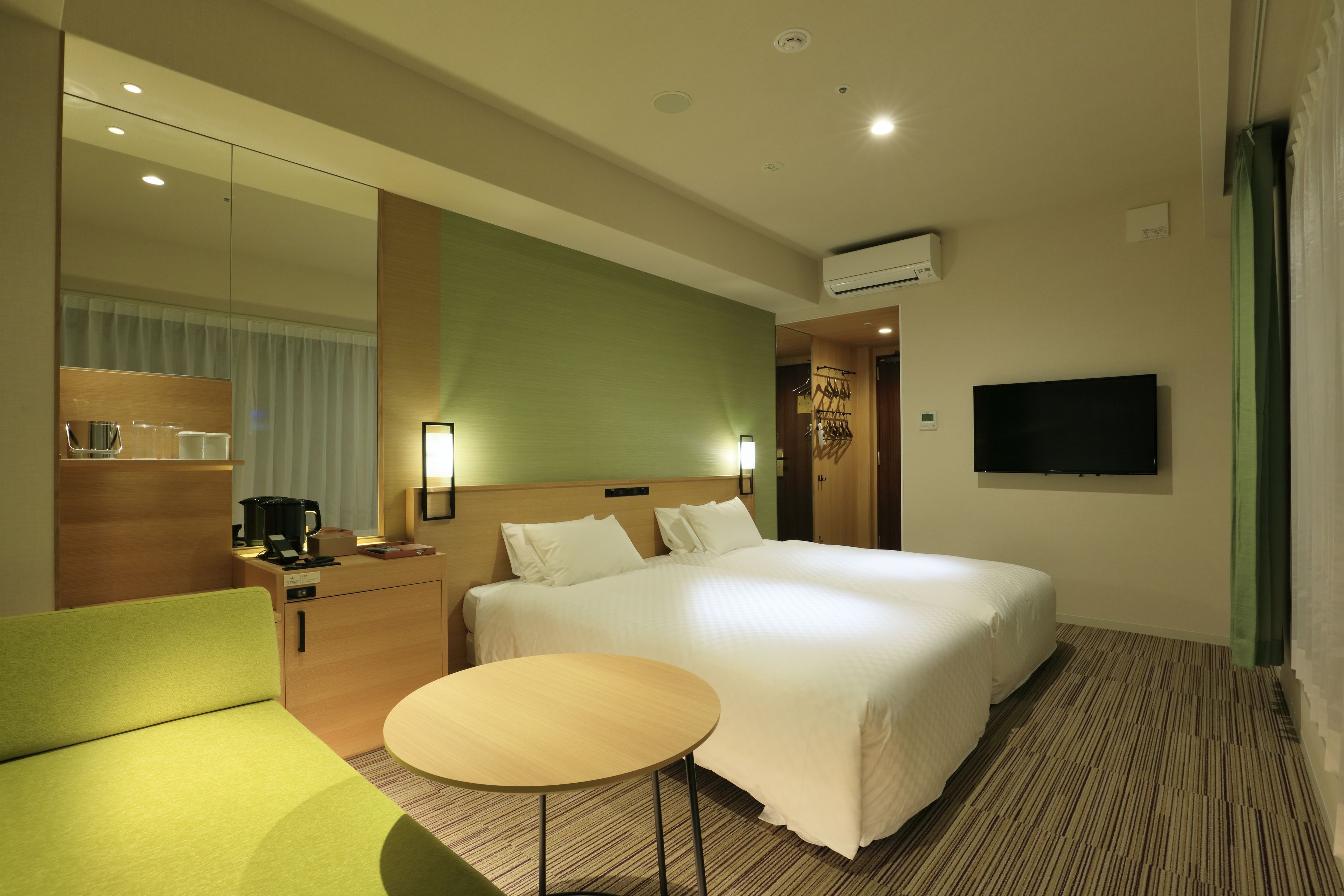 A hotel room with a large bed, a small table, a sofa, a TV, a coffee machine, and a mirror.
