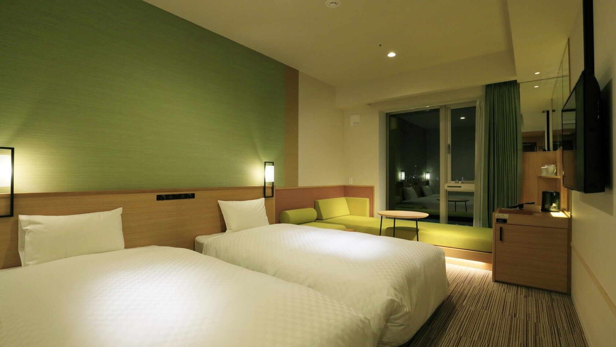 A hotel room with two beds, a sofa, a small table, a TV, and a view of the city at night.