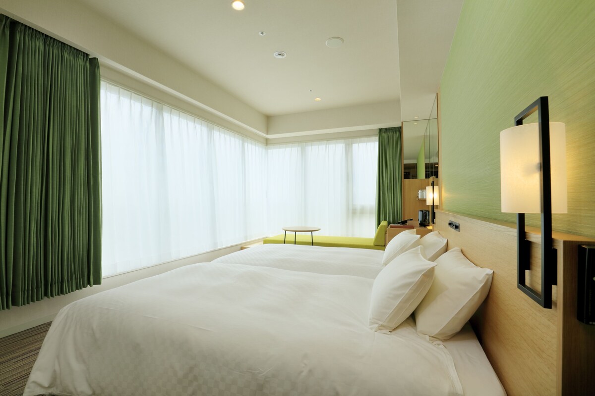 Habitación de hotel con dos camas, una mesita pequeña y un ventanal grande con cortinas.