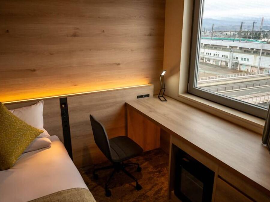 A hotel room with a bed, a desk, a chair, a lamp, and a window with a view of an industrial area.