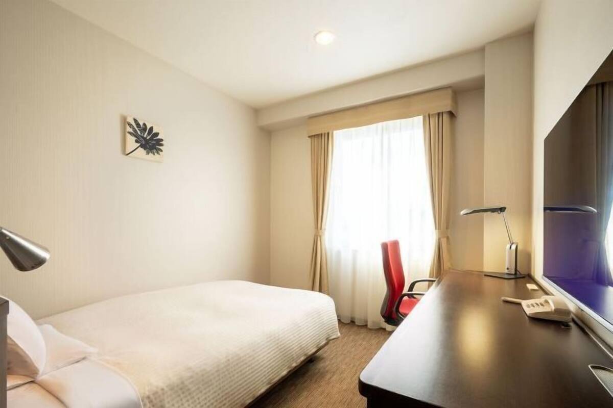 A hotel room with a bed, a desk, a lamp, a chair, and a TV.