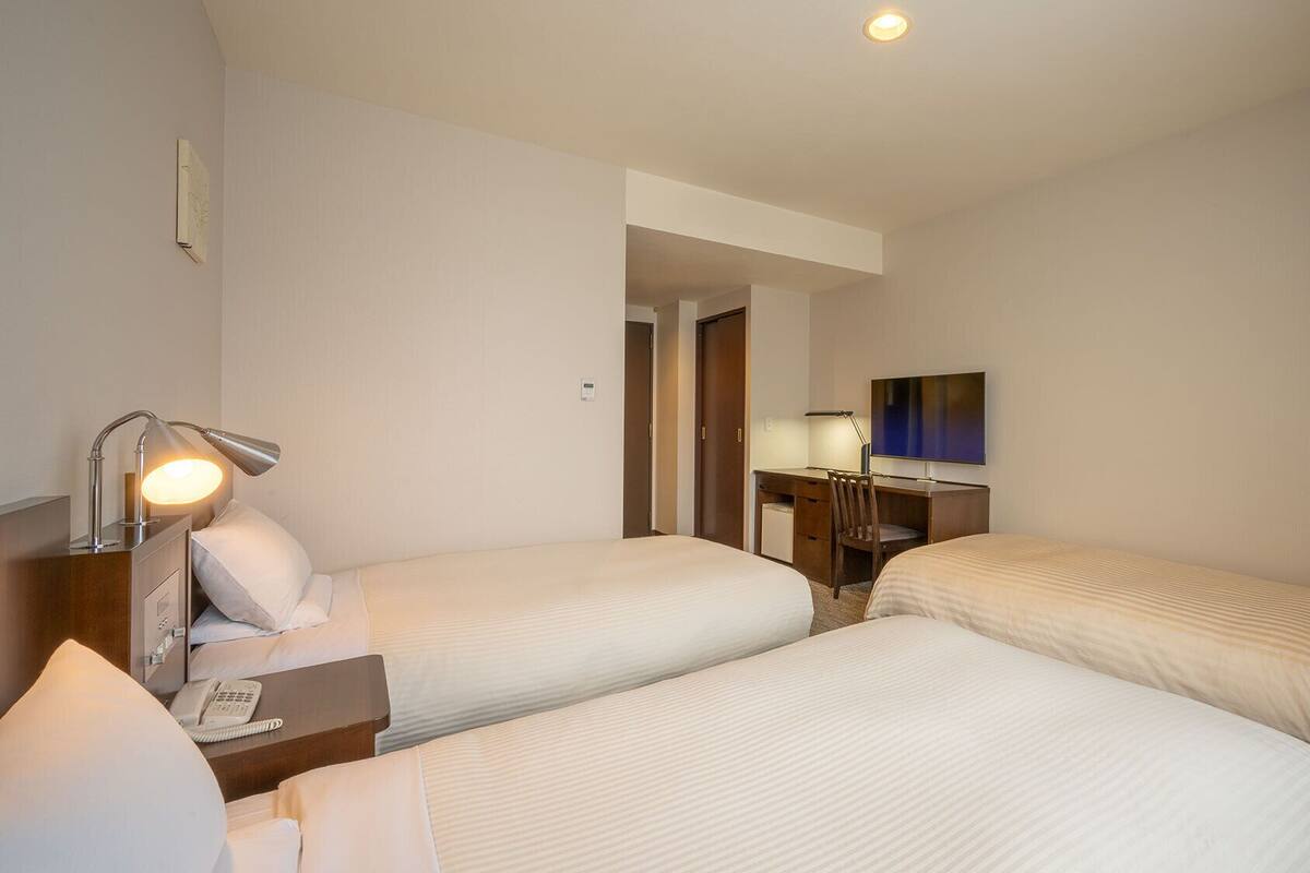 Ein Hotelzimmer mit zwei Betten, einem Schreibtisch mit Stuhl, einer Schreibtischlampe, einem Telefon und einem Flachbildfernseher.