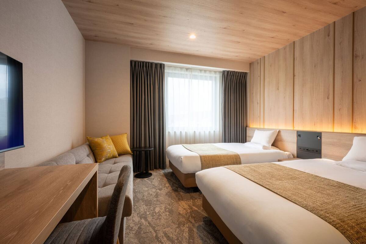 Ein Hotelzimmer mit zwei Betten, einem Holztisch, einem Stuhl, einem Fernseher und einem Fenster mit Vorhängen.