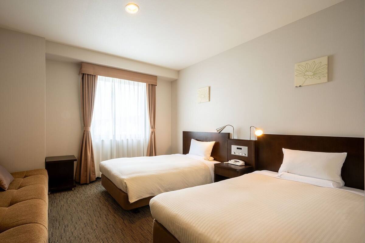 Ein Hotelzimmer mit zwei Betten, einem Schlafsofa, einem Nachttisch mit Lampe und einem Fenster mit Vorhängen.
