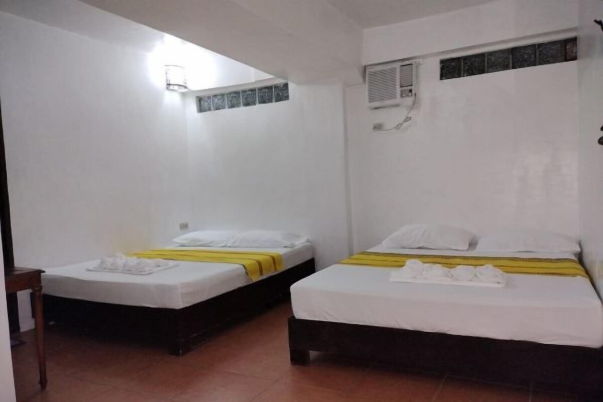 Ein Zimmer mit zwei Betten, jeweils mit weißer Bettwäsche und gelben Akzenten. Es gibt einen kleinen Tisch und eine wandmontierte Leuchte.