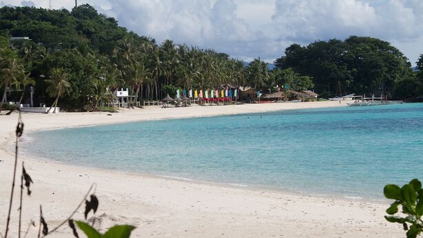 Une plage à l’eau turquoise et limpide, au sable blanc, et bordée d’une rangée de drapeaux aux couleurs vives.