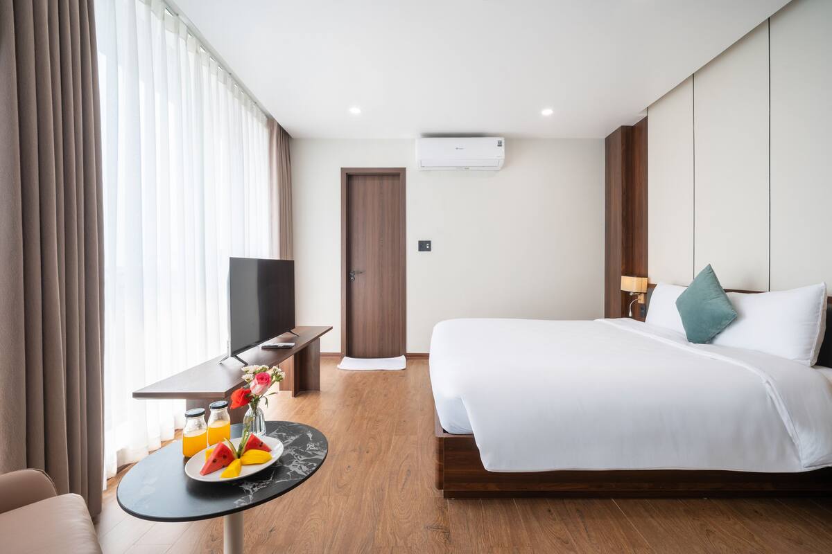 A hotel room with a bed, a desk with a laptop, a TV, and a breakfast setup with fruit and juice.