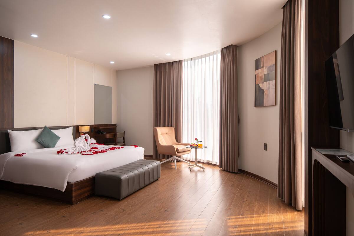 A hotel room with a large bed, a sitting area with a chair and table, a flat-screen TV, and a large window with curtains.