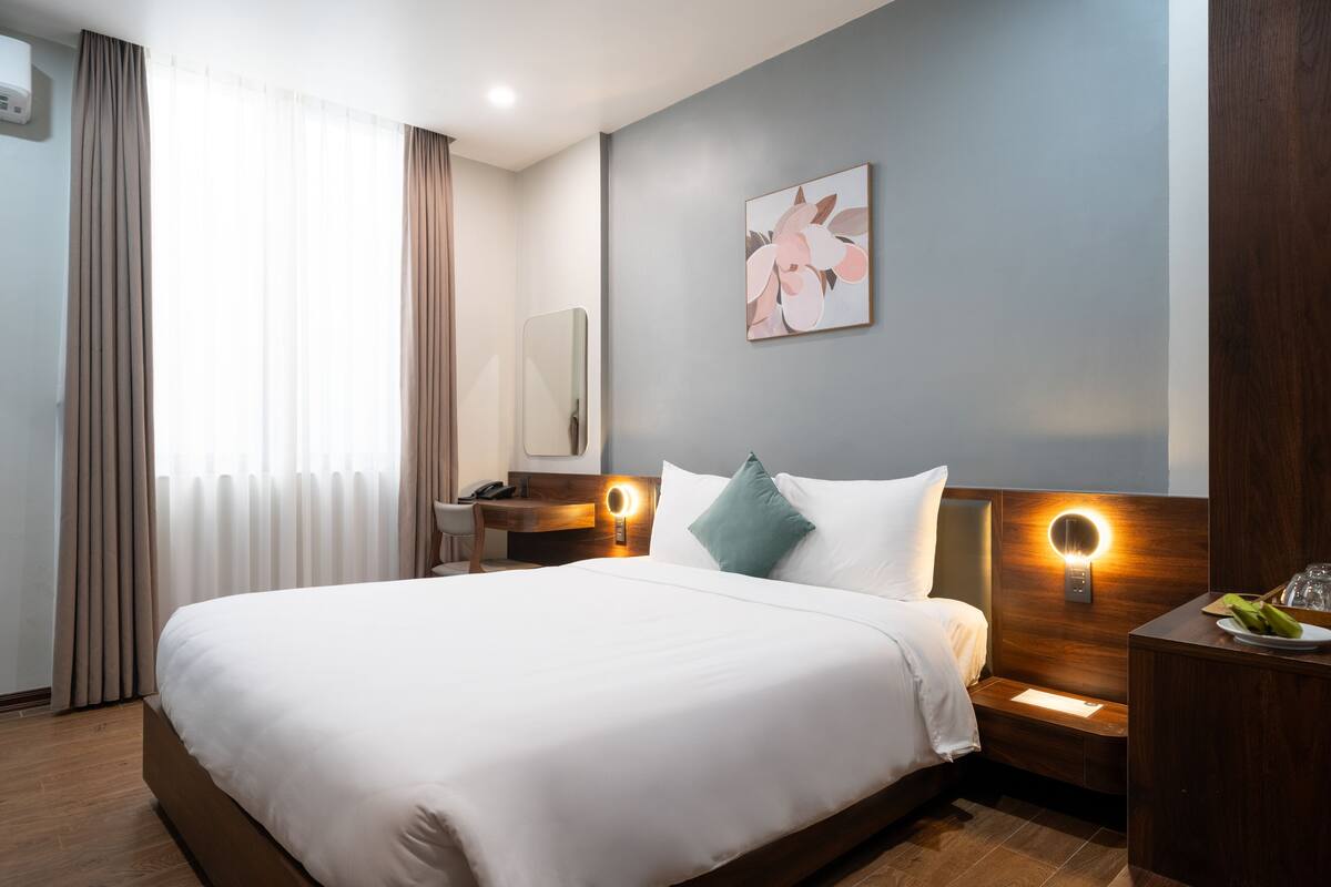 A modern hotel room with a large bed, a bedside table with a lamp, a mirror, and a wall-mounted light fixture.