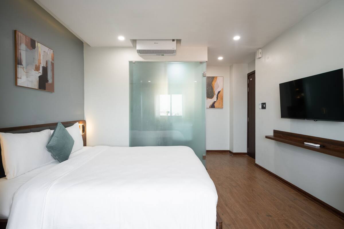 A hotel room with a bed, a TV, a glass door, and artwork on the walls.