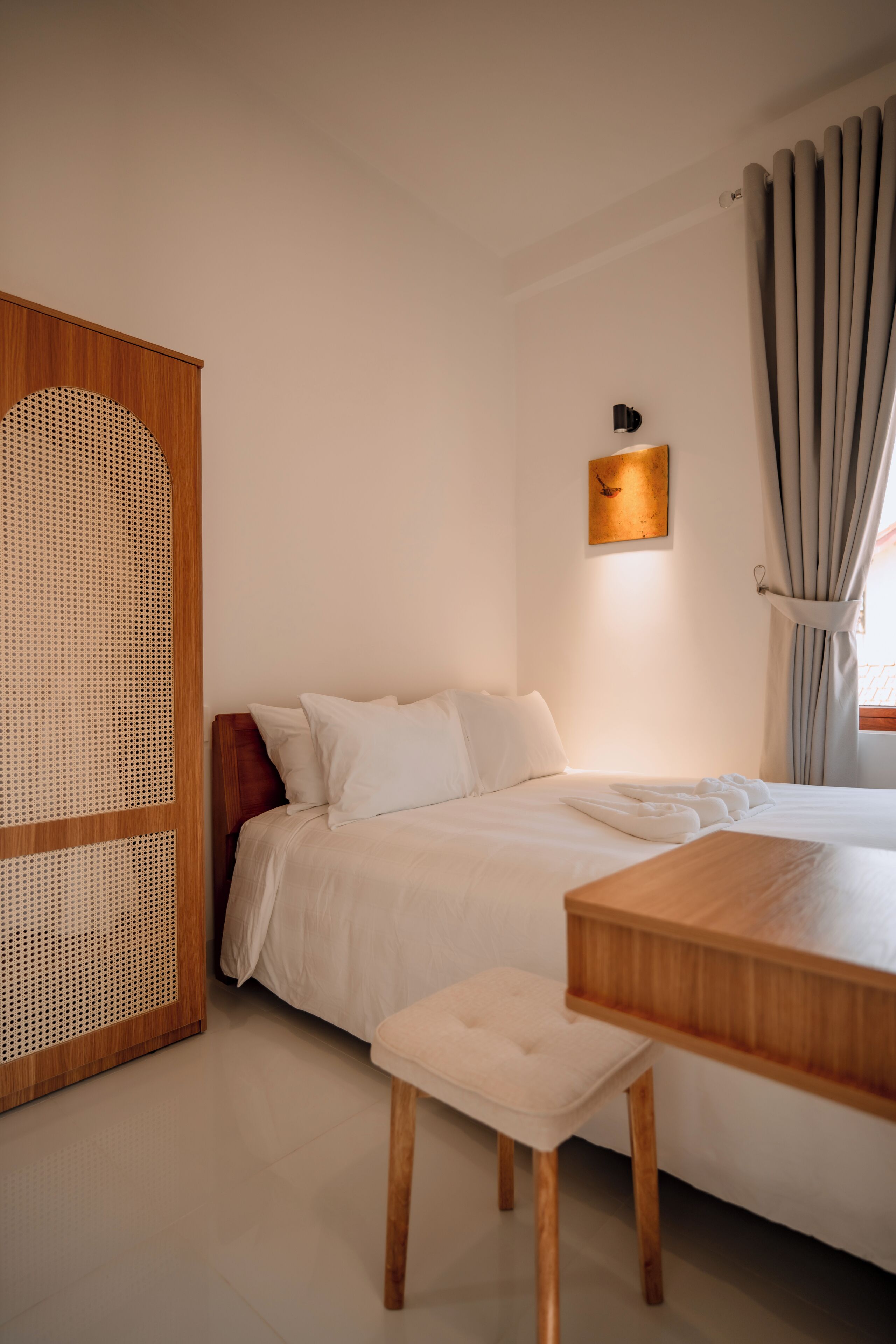 A neatly arranged bedroom with a bed, a wooden nightstand, a small stool, and a wall-mounted light fixture.