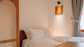 Un dormitorio ordenado con una cama, una mesita de noche de madera, un taburete pequeño y una lámpara de pared.
