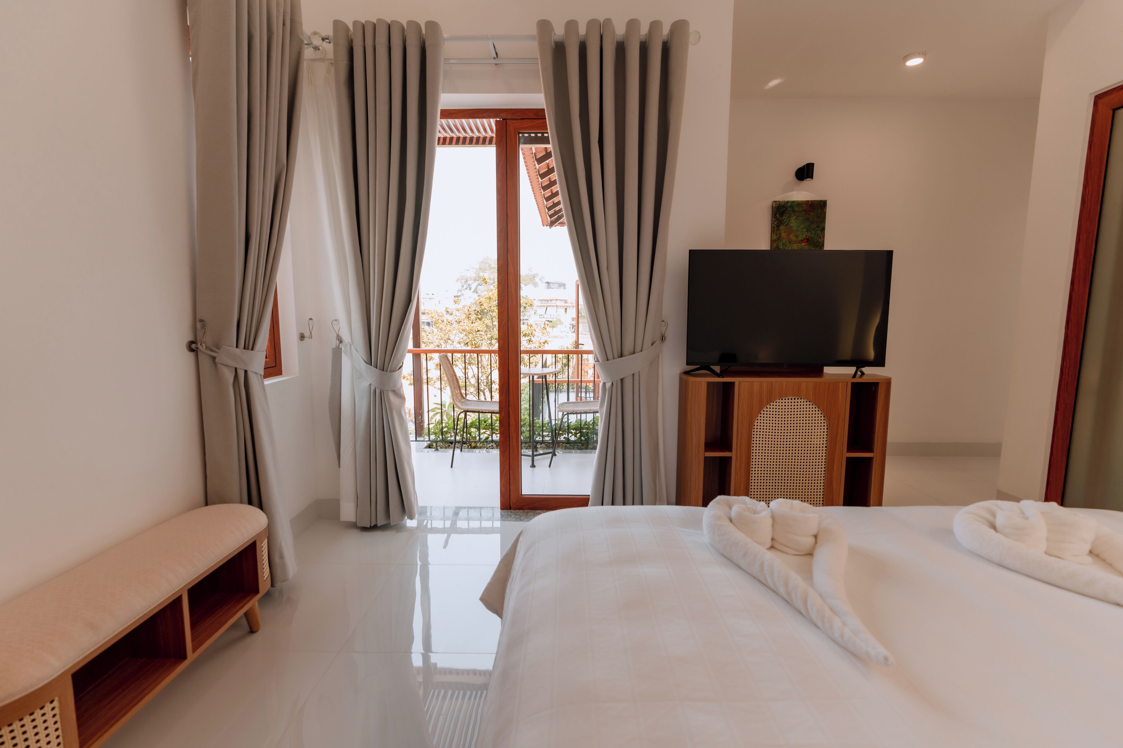 Habitación de hotel con cama, una banca, televisor sobre un mueble de madera y balcón con sillas.