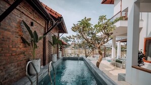 A swimming pool area with a brick wall, potted plants, and a view of a traditional building in the background. - Hue Riverside Villa (Hue)