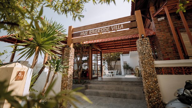 The entrance to HUE RIVERSIDE VILLA with a wooden archway, stone pillars, and a patio area with outdoor seating. - Hue Riverside Villa (Hue)