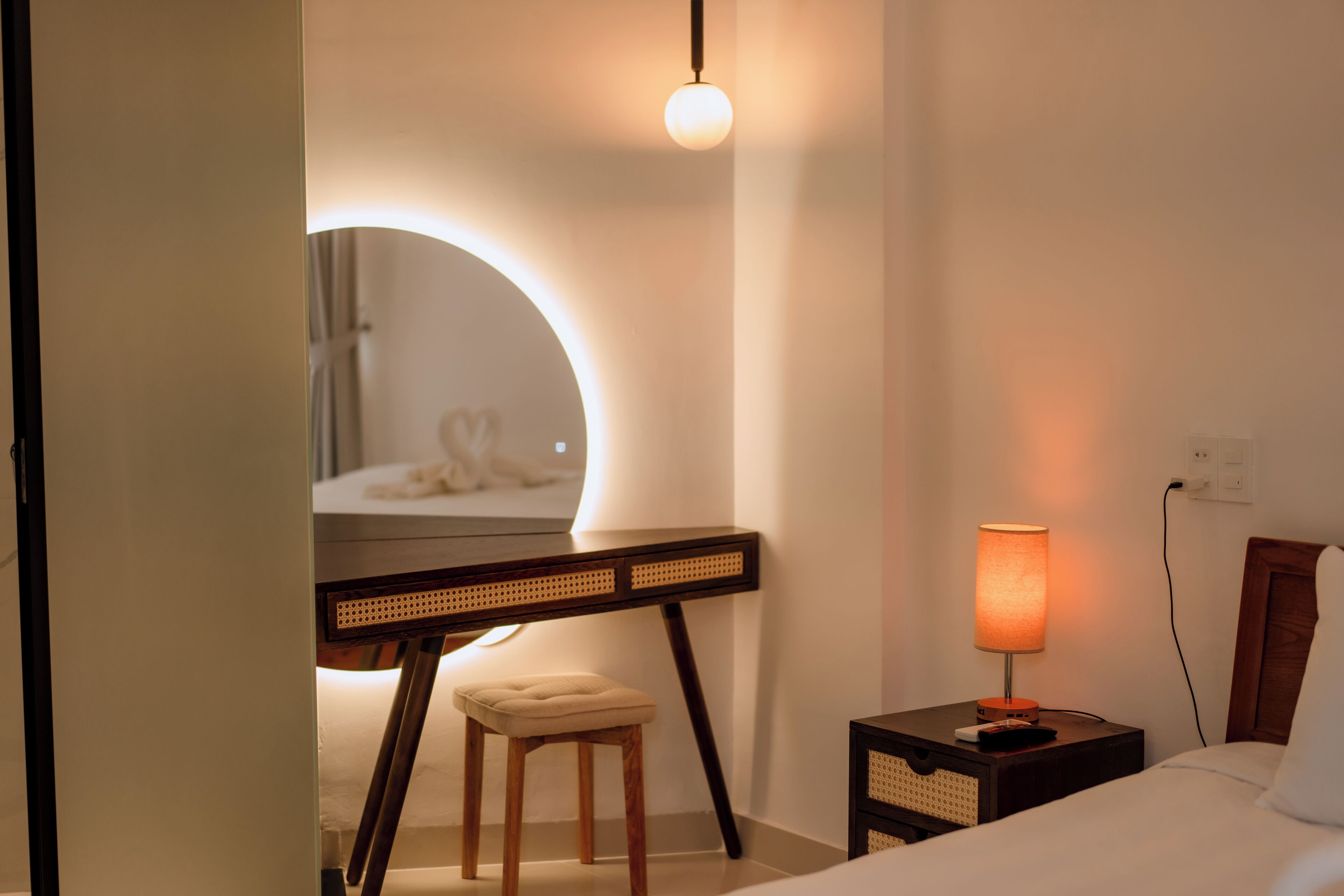 Habitación de hotel con cama, mesita de noche con lámpara, un taburete pequeño, escritorio con espejo y una lámpara circular.
