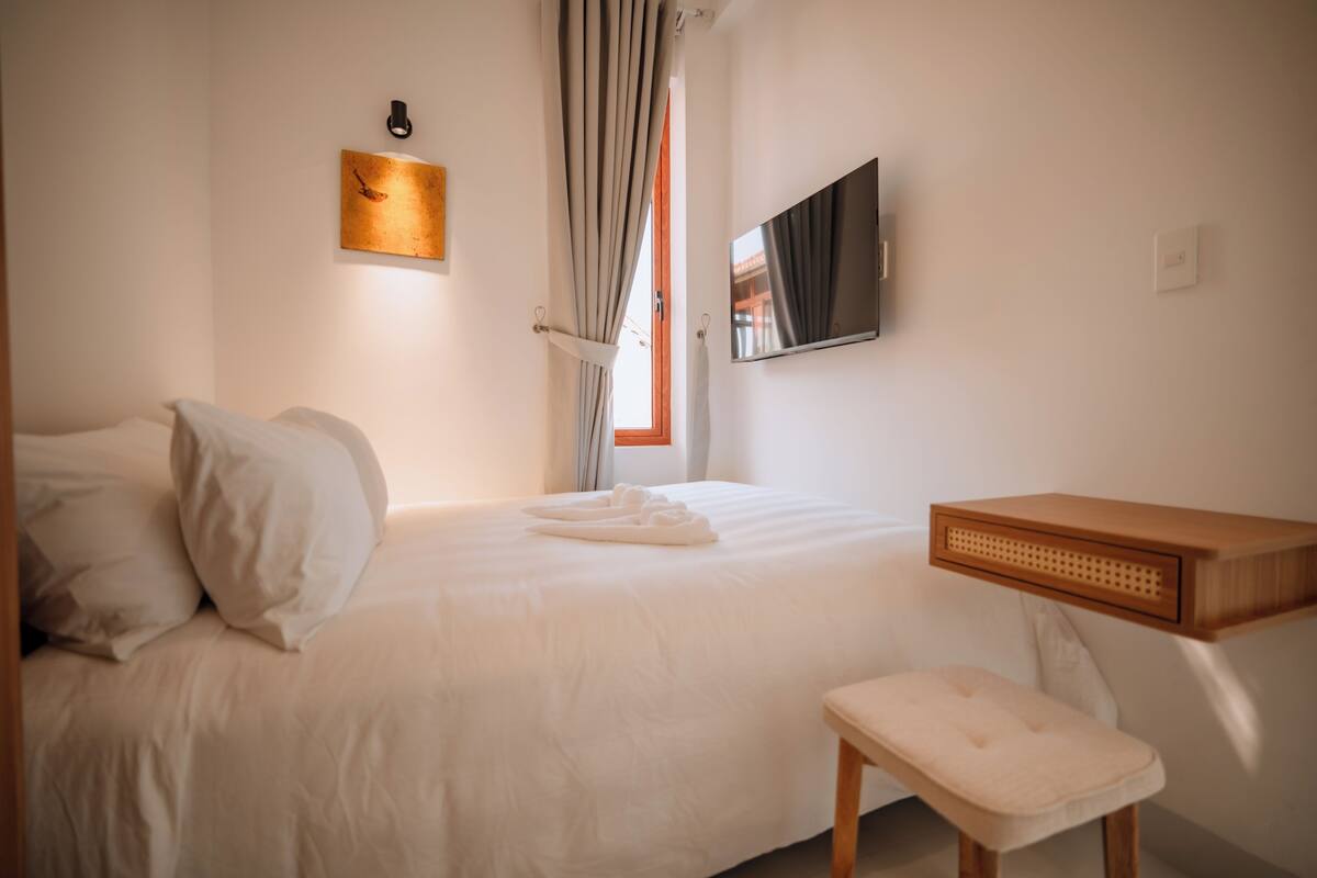 Quarto de hotel com cama, televisão, mesa de cabeceira de madeira e janela com cortinas.