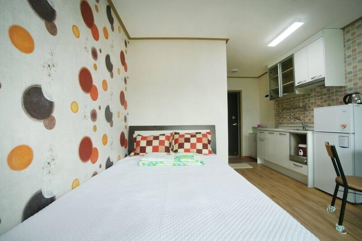 A bedroom with a bed, checkered pillows, a small kitchenette with a refrigerator, and a bathroom visible through an open door.