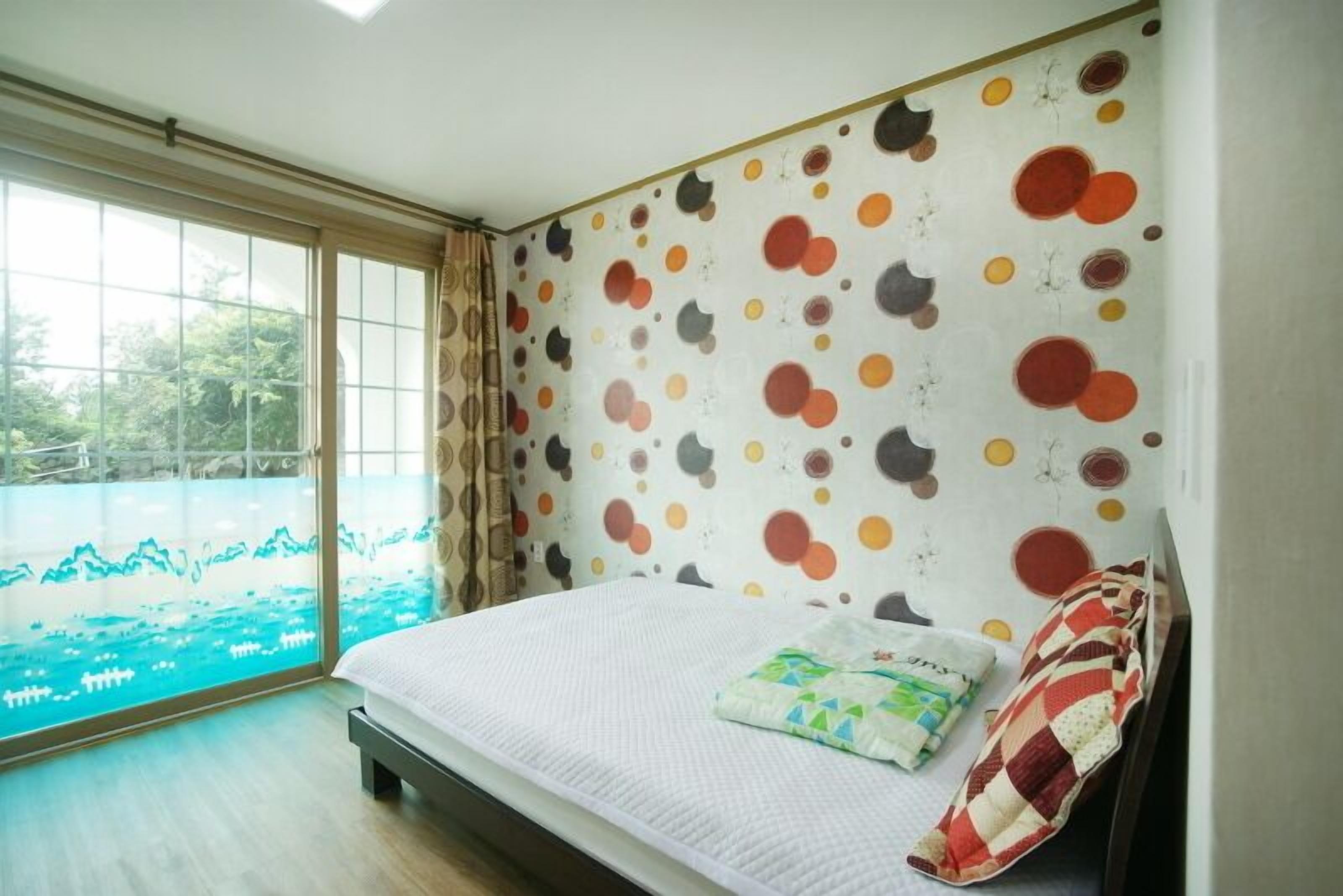 Une chambre avec une grande fenêtre, un lit doté d’un matelas blanc et d’un coussin coloré, et un mur orné d’un motif à pois.
