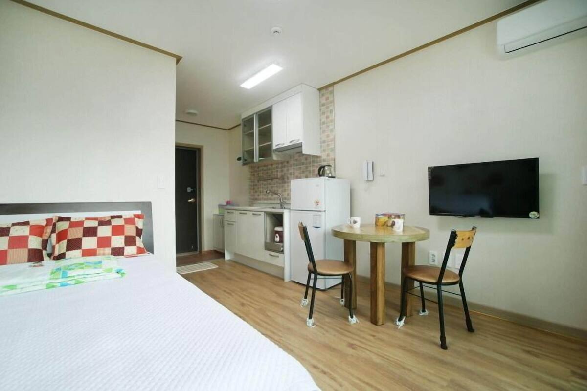 A compact room with a bed, a small kitchenette, a dining table with chairs, and a TV mounted on the wall.