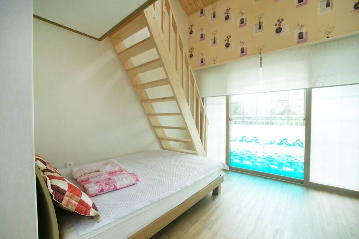 A room with a sloped ceiling, a wooden staircase, a bed with a quilt, and a large window with a view of a body of water.