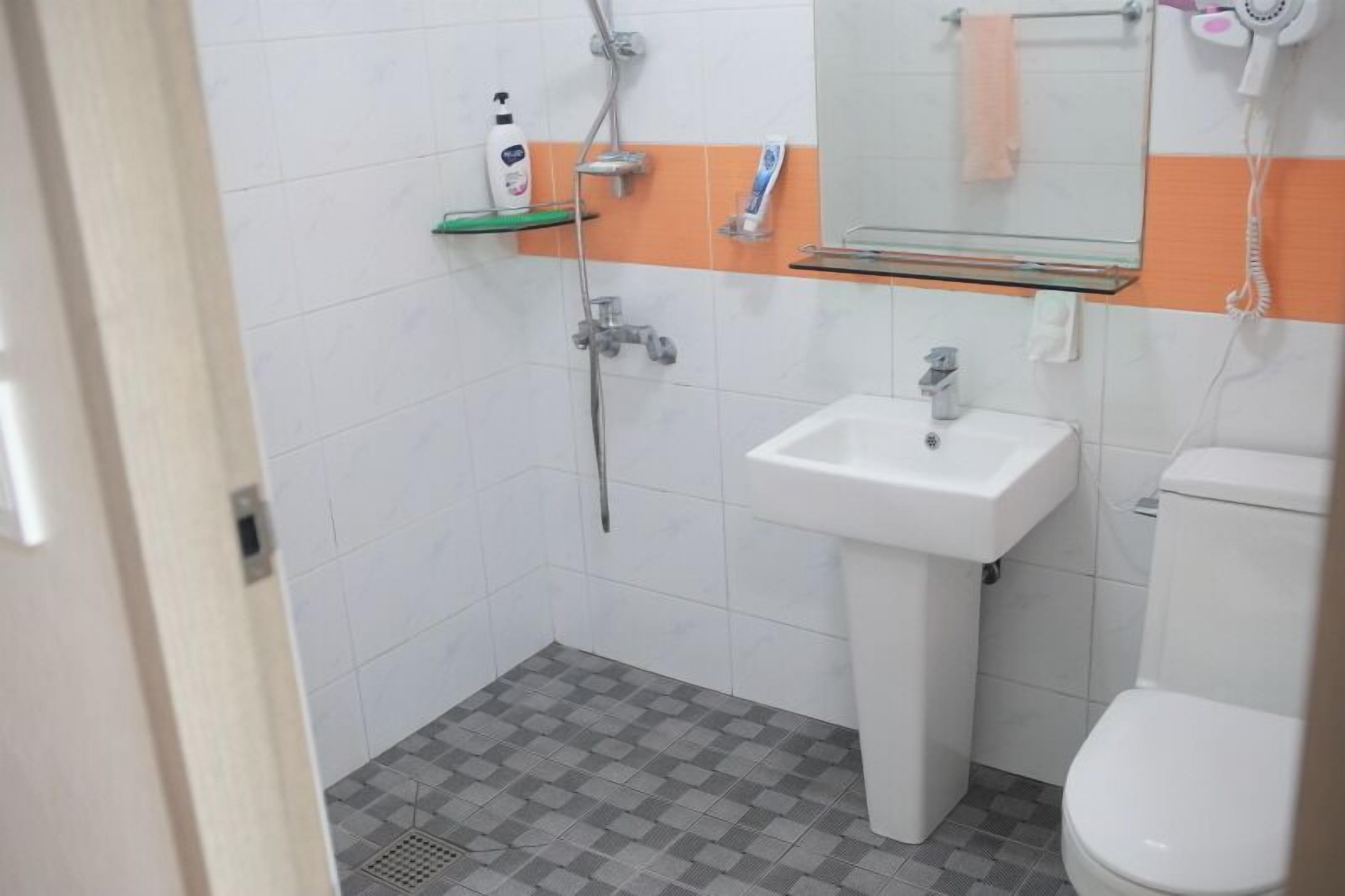 Une salle de bain avec un lavabo blanc, des toilettes et une douche. On y trouve une étagère en verre avec des articles de toilette et un porte-serviettes.