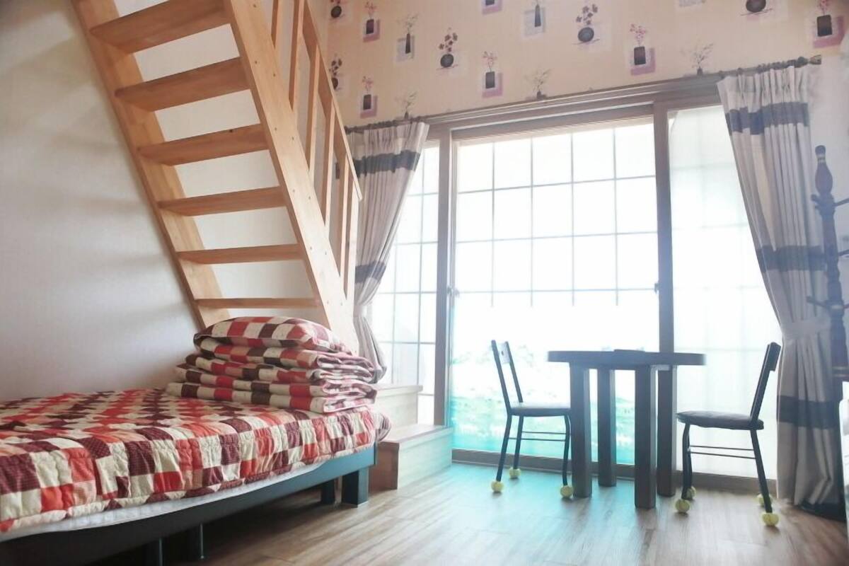 A bedroom with a bed, a wooden ladder, a table with chairs, and a large window with curtains.