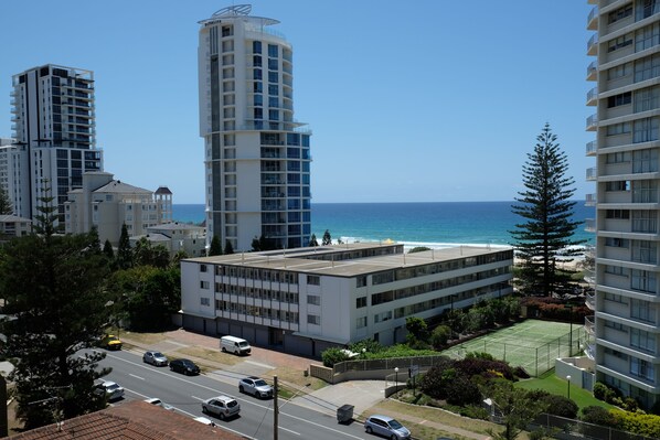A high-rise building with a tennis court in the foreground, surrounded by other buildings and a clear view of the ocean.