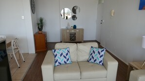 A living room with a beige sofa, two patterned cushions, a wooden cabinet, and a small table with a lamp.