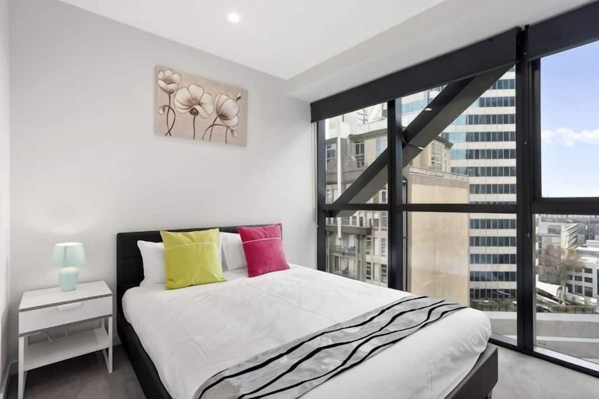 Quarto moderno com uma cama grande, uma mesa de cabeceira, uma luminária e vista da cidade através de janelas amplas.