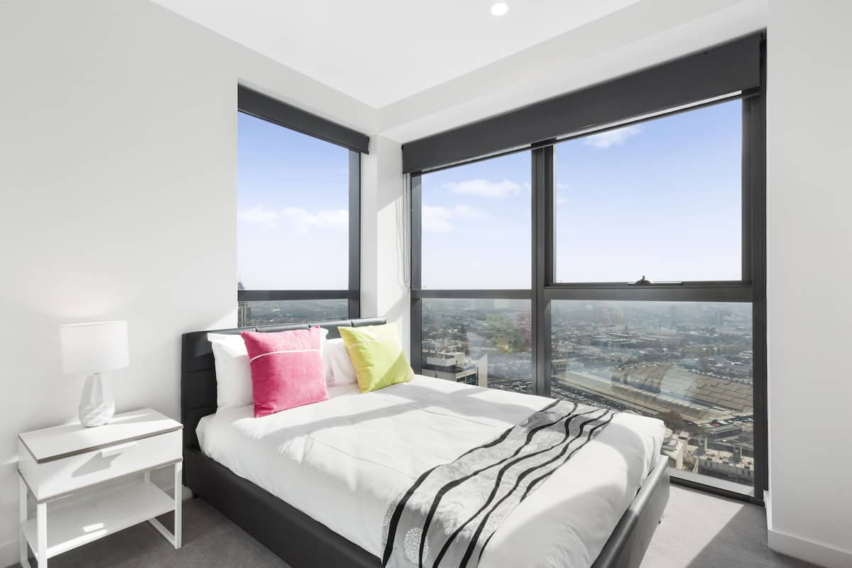 Quarto moderno com uma cama grande, mesa de cabeceira e vista para a cidade através de amplas janelas.